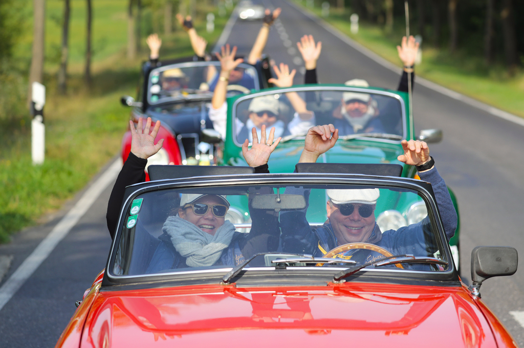 Frontalaufnahme von drei Oldtimer-Cabriolets, die eine Allee entlangfahren. Die Insassen reißen gut gelaunt die Arme in die Luft.