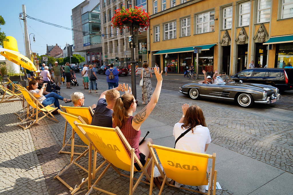 ADAC Landpartie Classic 2019 ⓒ Kai-Uwe Knoth #3415 Ein begeistertes Publikum sitzt in gelben Liegestühlen am Straßenrand und winkt einem vorbeifahrenden Oldtimer-Cabrio zu. Es ist sonnig, im Hintergrund sind weitere Zuschauerinnen, ein gelber ADAC-Sonnenschirm und historische Gebäude zu sehen.