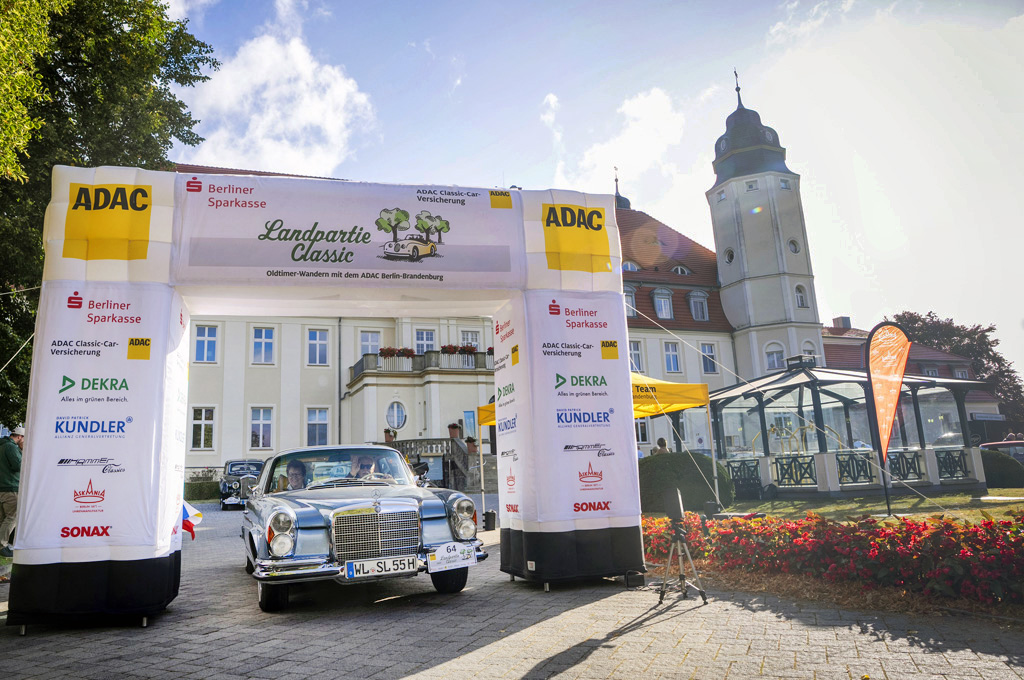 Die Sonne scheint. Ein silberner Mercedes-Oldtimer fährt durch ein weißes Starttor der „ADAC Landpartie Classic“ vor dem Schloss Fleesensee. Auf dem Tor sind Sponsorenlogos zu sehen, daneben Blumenbeete.