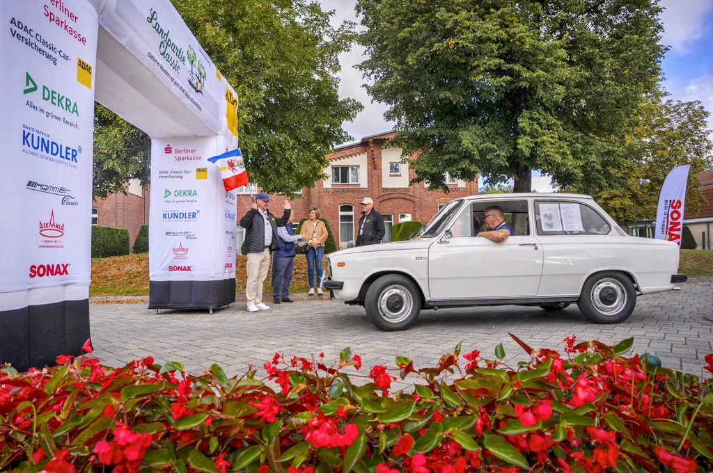 Ein weißer Oldtimer fährt unter einem Torbogen mit Sponsorenlogos hindurch. Daneben stehen mehrere Personen, die dem Fahrer zuwinken. Im Vordergrund sind rote Blumen, im Hintergrund ein Backsteingebäude und Bäume.