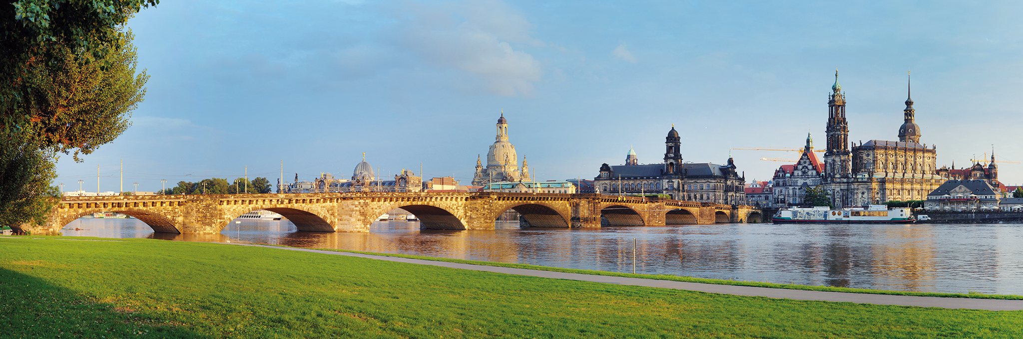 Panoramablick über die Elbe auf die Dresdner Altstadt im warmen Abendlicht: Die Augustusbrücke aus Sandstein spannt sich über den Fluss, dahinter ragen die Kuppel der Frauenkirche sowie die Türme von Ständehaus und Hofkirche auf, im Vordergrund liegt eine grüne Uferwiese.