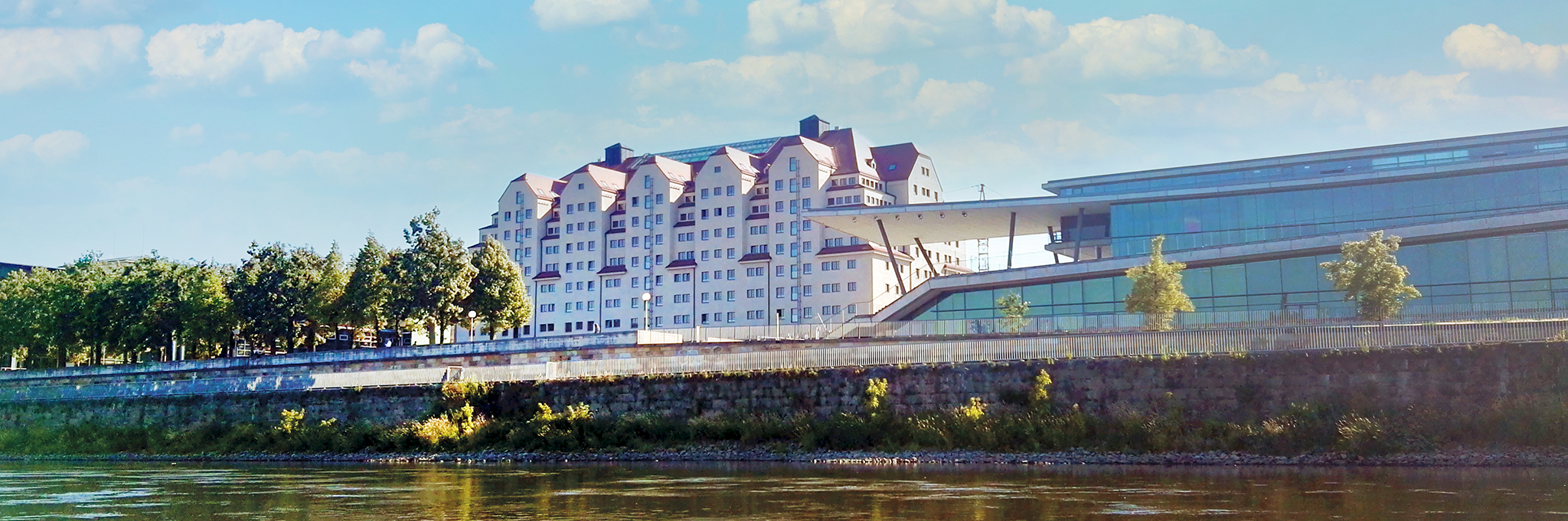 Blick von der Elbe auf das Ufer in Dresden: Das Maritim Hotel Dresden mit stufenförmigen Giebeln und roten Dächern steht neben einem modernen Glasbau mit weit auskragendem Dach, davor eine Ufermauer mit Grün, darüber ein blauer Himmel mit Wolken.
