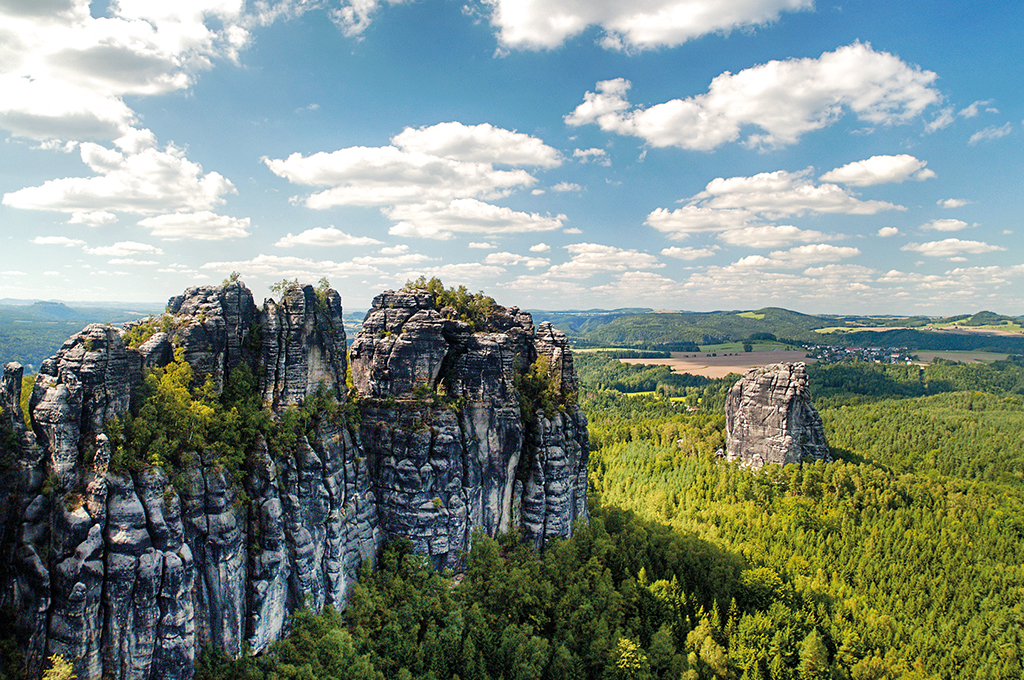 Felsformationen der Schrammsteine in der Sächsischen Schweiz: Links ragen zerklüftete, bewachsene Sandsteinfelsen auf, rechts steht ein einzelner Felsblock über einem dichten grünen Wald, dahinter erstreckt sich eine weite Hügellandschaft unter blauem Himmel mit Wolken.