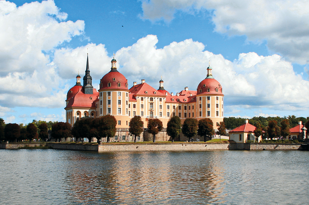 Schloss Moritzburg bei Dresden: Das gelb-ockerfarbene Barockschloss mit roten Kuppeln und Dächern liegt auf einer Insel im Schlossteich, davor spiegeln sich Bäume und Himmel im Wasser, darüber türmen sich große weiße Wolken am blauen Himmel.