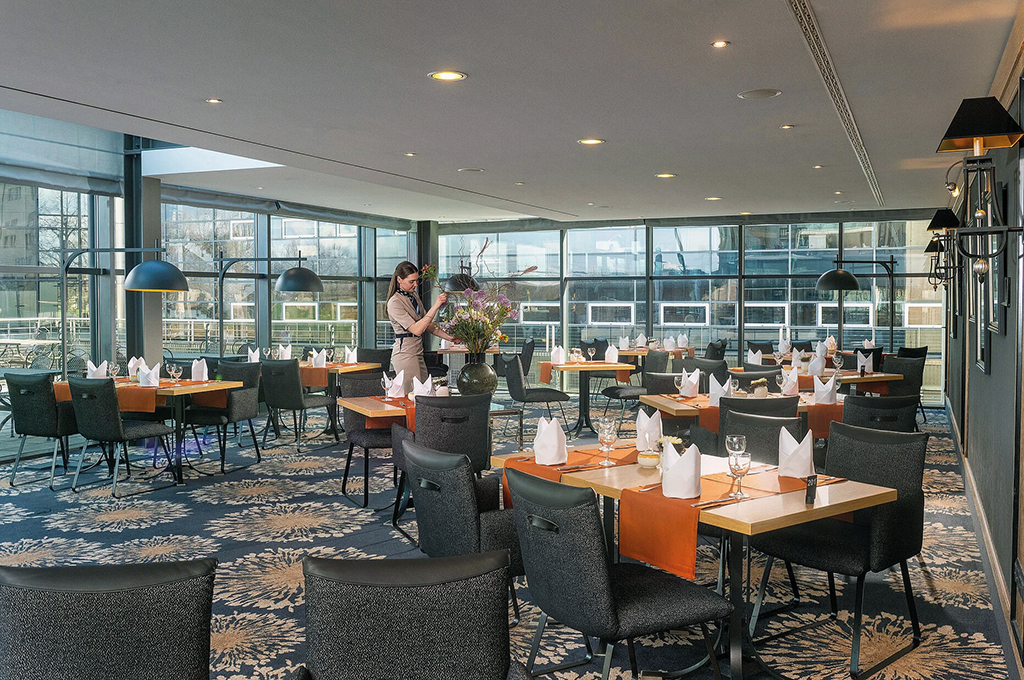 Restaurant des Maritim Hotel Dresden mit Fensterfront: Eingedeckte Tische mit gefalteten weißen Servietten und orangefarbenen Läufern stehen auf blau gemustertem Teppich, im Hintergrund arrangiert eine Mitarbeiterin einen Blumenstrauß.