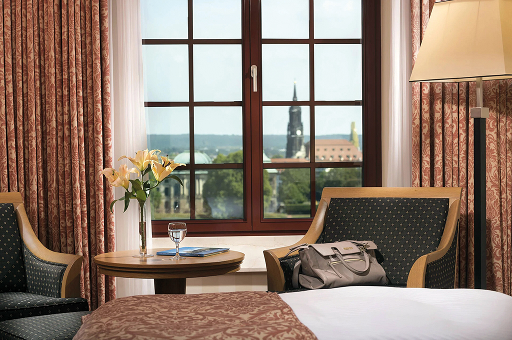 Superior-Hotelzimmer im Maritim Hotel Dresden mit Blick auf Dresden: Zwei gepolsterte Sessel mit Holzrahmen und ein runder Tisch mit Lilien und Wasserglas stehen vor einem Sprossenfenster, durch das der Turm der Kreuzkirche und die Dächer der Altstadt zu sehen sind.